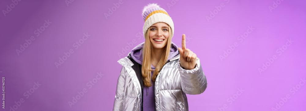 Wall mural Excited cheerful fair-haired european girl in winter hat silver shiny jacket show number one index fingers give suggestion advice smiling broadly talking happily, purple background