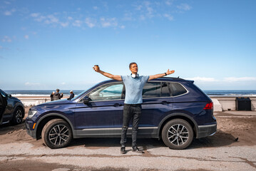 Fototapeta na wymiar San Francisco, USA. September 20, 2022. Carefree mid adult man with arms outstretched standing by blue car at beach with sea and sky in the background during summer