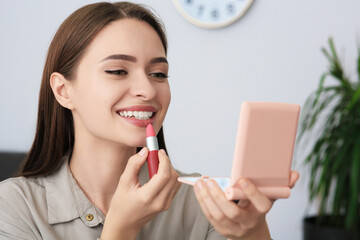 Young woman with cosmetic pocket mirror applying lipstick indoors