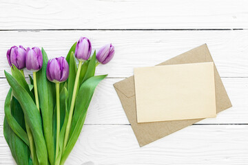 Bouquet of purple tulips and blank greeting card on white wooden background, top view, copy space.