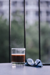 Hot coffee cup with eye glasses on table against balcony. Close up