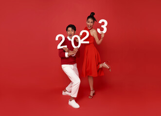 Happy asian couple in red casual attire showing number 2023 greeting happy new year with smiles on bright red background.