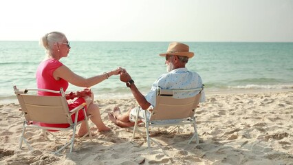 Slow motion. Elderly couple man and woman wearing fashion sunglasses talking together and looking at the sea sky sitting on chair on beach. Vacation trip summer holiday. - Powered by Adobe