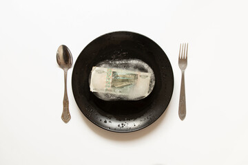 Ten rubles in a frozen block of ice lie on a plate next to a spoon and fork, Russian finance and...