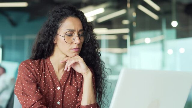 Young serious concerned hispanic businesswoman sitting at desk looking laptop computer in contemporary corporation office Business technologies concept portrait thoughtful freelancer employee
