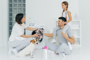 Indoor shot of affectionate father plays with daughter in washing room, gives checkered dirty shirt to wife, pose together near washer, surrounded with washing powder bottle, white furniture
