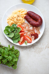 Breakfast with scramble, bacon, chorizo, tomatoes and fresh greens on a white plate, above view on a light-beige stone background, vertical shot with space