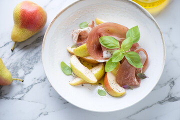 Prosciutto with pear wedges and fresh basil served in a beige plate, elevated view on a white marble background