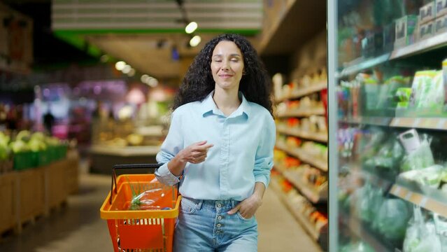young hispanic woman shopping in a supermarket walking and choosing products in a grocery store. Happy female shopper with basket between shelves doing daily shopping Browses through Goods Section