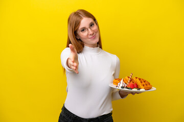 Young redhead woman holding waffles isolated on yellow background shaking hands for closing a good deal