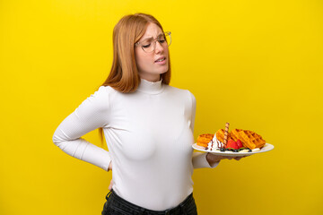 Young redhead woman holding waffles isolated on yellow background suffering from backache for having made an effort