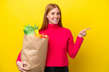 Young redhead woman holding a grocery shopping bag isolated on yellow background pointing finger to...