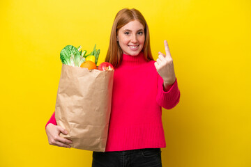 Young redhead woman holding a grocery shopping bag isolated on yellow background doing coming...