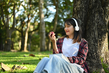 Happy young asian woman eating apple under huge tree on green grass at sunny beautiful garden background