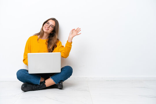 Young Caucasian Woman With A Laptop Sitting On The Floor Isolated On White Background Saluting With Hand With Happy Expression