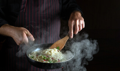 Chef prepares delicious spaghetti with spices and seasonings in a steamed hot frying pan. Space for advertising on a dark background. Menu for a restaurant or hotel
