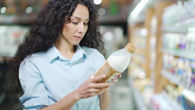 Hispanic woman chooses dairy products holding a bottle of milk in her hands. female stands in a grocery store picking shopping Studies and reads information on the label. Considers makes choices