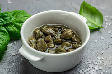 Marinated capers with fresh basil leaves in a small bowl, on light wooden background. Food recipe background. Close up
