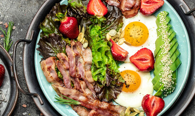 Two eggs, bacon, avocado, strawberries and fresh salad for healthy breakfast on a dark background....