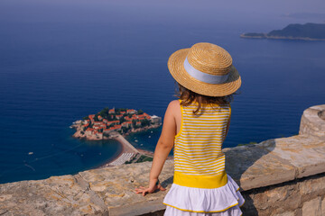Rear view of little girl with straw hat looking island of Sveti Stefan with hotel resort in Adriatic sea, Budva, Montenegro