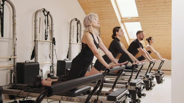 Slim good looking senior woman doing core exercises while taking pilates lesson on a reformer machine, by female instructor at a big gymnasium center, wide industrial studio. Healthy fitness lifestyle