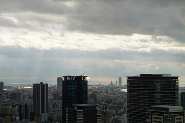 Overhead view of Osaka's Umeda area from a hill on a cloudy day, sunlight shining through a gap in the clouds.