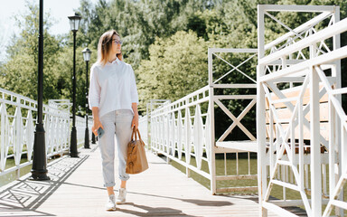 Stylish young female student wearing glasses, bag and holding smartphone walking in park on sunny day and looking away