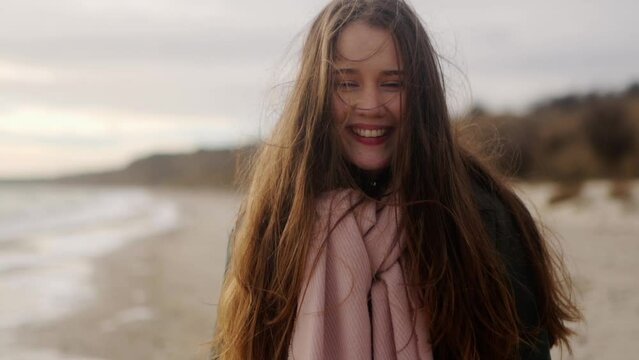 Happy mixed ethnic girl smiling and looking at camera on sea shore. Portrait of loving joyful asian woman laughing, standing on ocean coast. Wind blows her airy hair. Dreamy cinematic slow motion.
