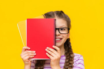 A beautiful little girl looks out from behind a red notebook and smiles. A schoolgirl on a yellow isolated background.