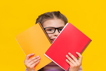 A beautiful little girl looks out from behind a red notebook and smiles. A schoolgirl on a yellow isolated background.