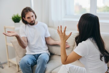 A man and a woman sit on the couch at home in white T-shirts and chatting scandalously do not understand each other. A quarrel in the family of two spouses and aggression