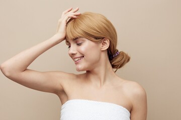 a sweet, elegant woman stands on a beige background wrapped in a white towel and smiling pleasantly, holding her hand on her head touching her hair, turning her face away with her eyes closed