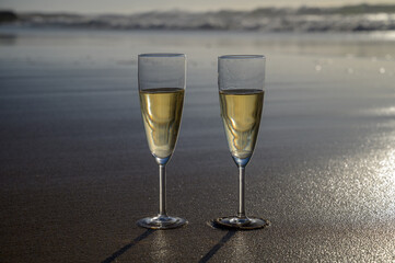 Glasses of cava or champagne sparkling wine on white sandy ocean beach with water waves on sunset in sunlights
