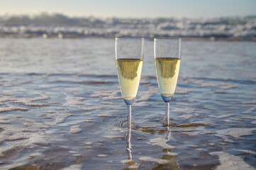Glasses of cava or champagne sparkling wine on white sandy ocean beach with water waves on sunset in sunlights