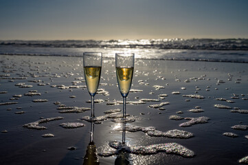 Glasses of cava or champagne sparkling wine on white sandy ocean beach with water waves on sunset...