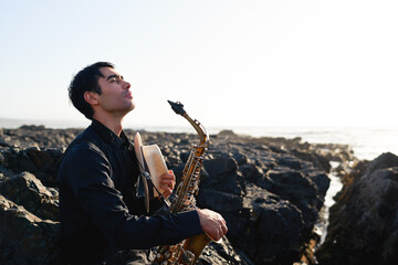 young latino saxophonist sitting on the rocks on the beach relaxed with eyes closed in a moment of...