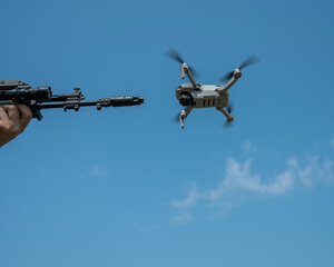 A man aims to shoot a rifle at a flying drone against a blue sky. 