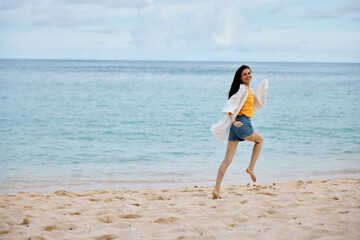 Fototapeta premium Sports woman runs along the beach in summer clothes on the sand in a yellow T-shirt and denim shorts white shirt flying hair ocean view, beach vacation