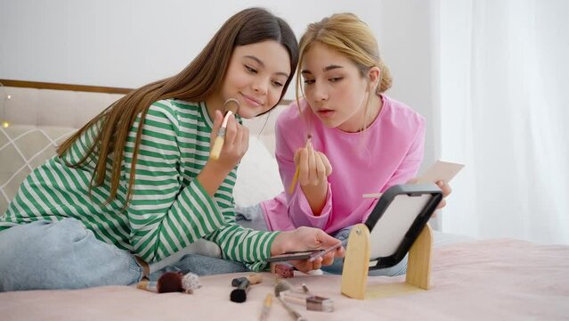 Beautiful adolescent girls, close friend, sitting on the bed, studying their first makeup at home