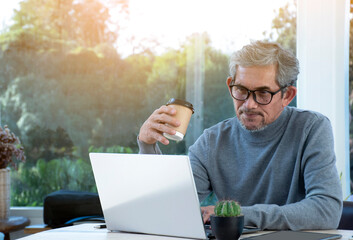Portrait elderly asian business man sits near transparent glass window and looking down to laptop on table in the morning of the day, sunlight edited, soft and selective focus, happy senior concept.