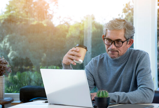 Portrait elderly asian business man sits near transparent glass window and looking down to laptop on table in the morning of the day, sunlight edited, soft and selective focus, happy senior concept.