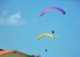 two colorful paragliders gliding over houses under a blue sky