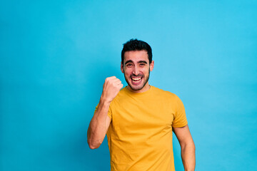 Handsome bearded male in yellow shirt raising arms and looking at camera while standing against blue background