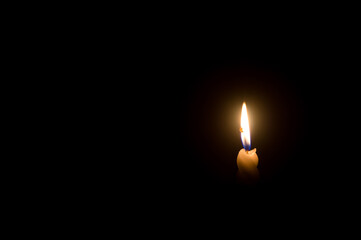 A single burning candle flame or light glowing on a spiral white candle on black or dark background on table in church for Christmas, funeral or memorial service