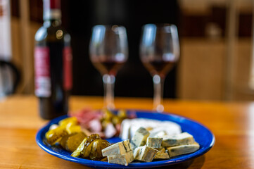 A plate filled with different kinds of cheese, olives, peppers, sausage, garlic and cucumber and a bottle of red wine with two glasses.