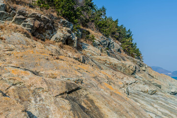Sloping rocky cliffs topped with evergreen trees under blue sky.