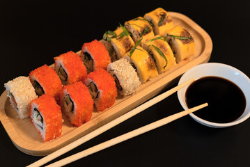 Delicious Japanese rolls with salmon, avocado, cucumber, cheese on wooden plate with wood sticks and soy sauce with selective focus on black background. Asian food concept. Healthy eating