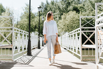Fototapeta na wymiar Sad young student woman with bag and phone wearily walking along bridge in park and looking away
