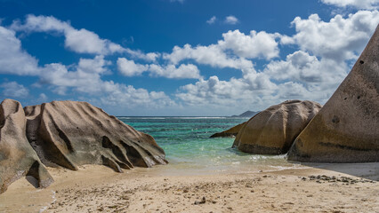 Picturesque granite rocks with rounded folded slopes on the beach of a tropical island. The waves of the turquoise ocean spread over the sand. Clouds in the blue sky. Seychelles. La Digue. 