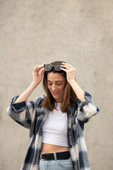 Caucasian girl in jeans and black and white plaid shirt looking to the side with sunglasses on her head, urban gray concrete background